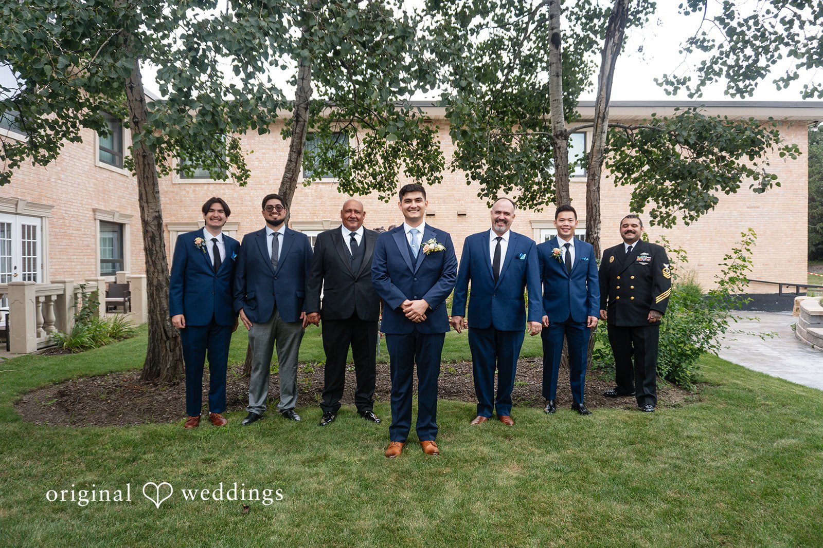 Selena + Ezequiel A group of men in matching dark blue suits pose standing together in a park.
