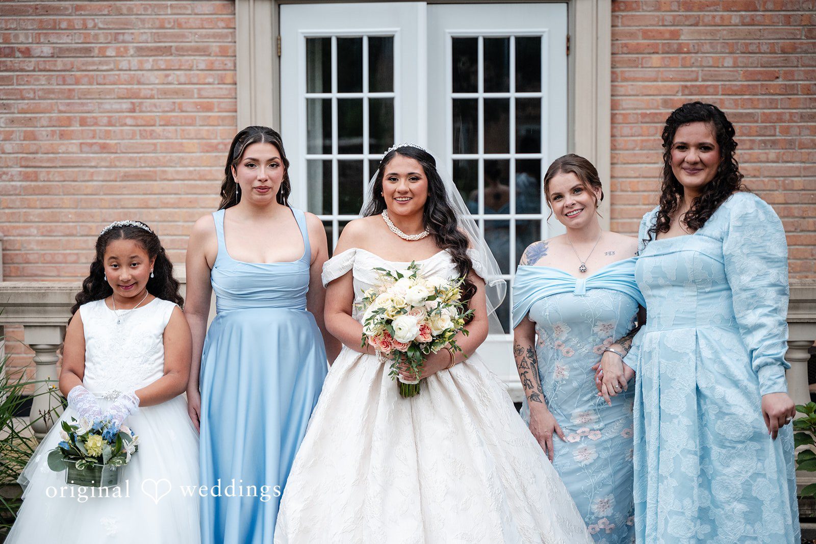 Selena + Ezequiel A bride stands with bridesmaids in matching light-blue dresses in front of a white door.