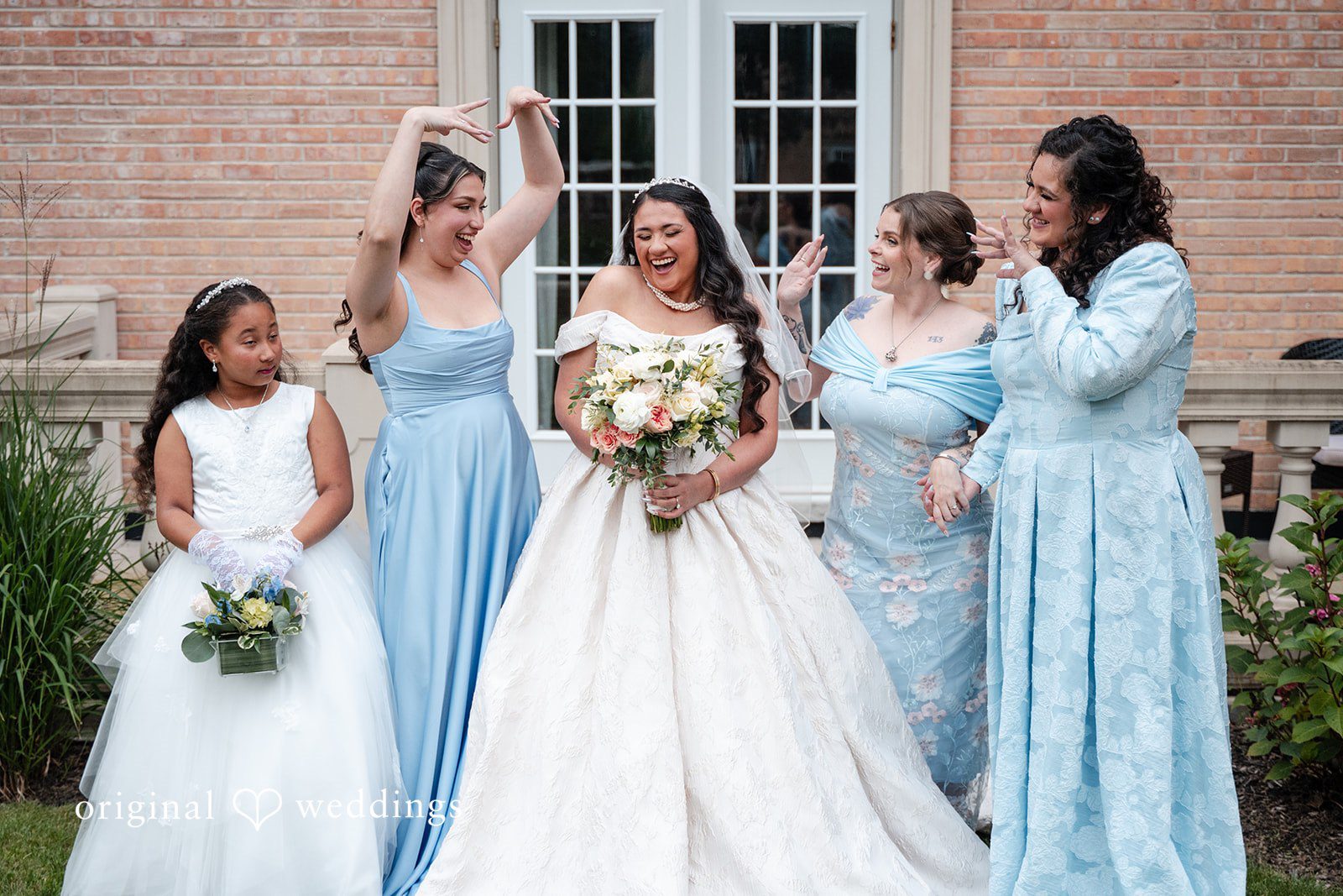 Selena + Ezequiel A bridal party in light-blue dresses poses together holding bouquets in front of a building.