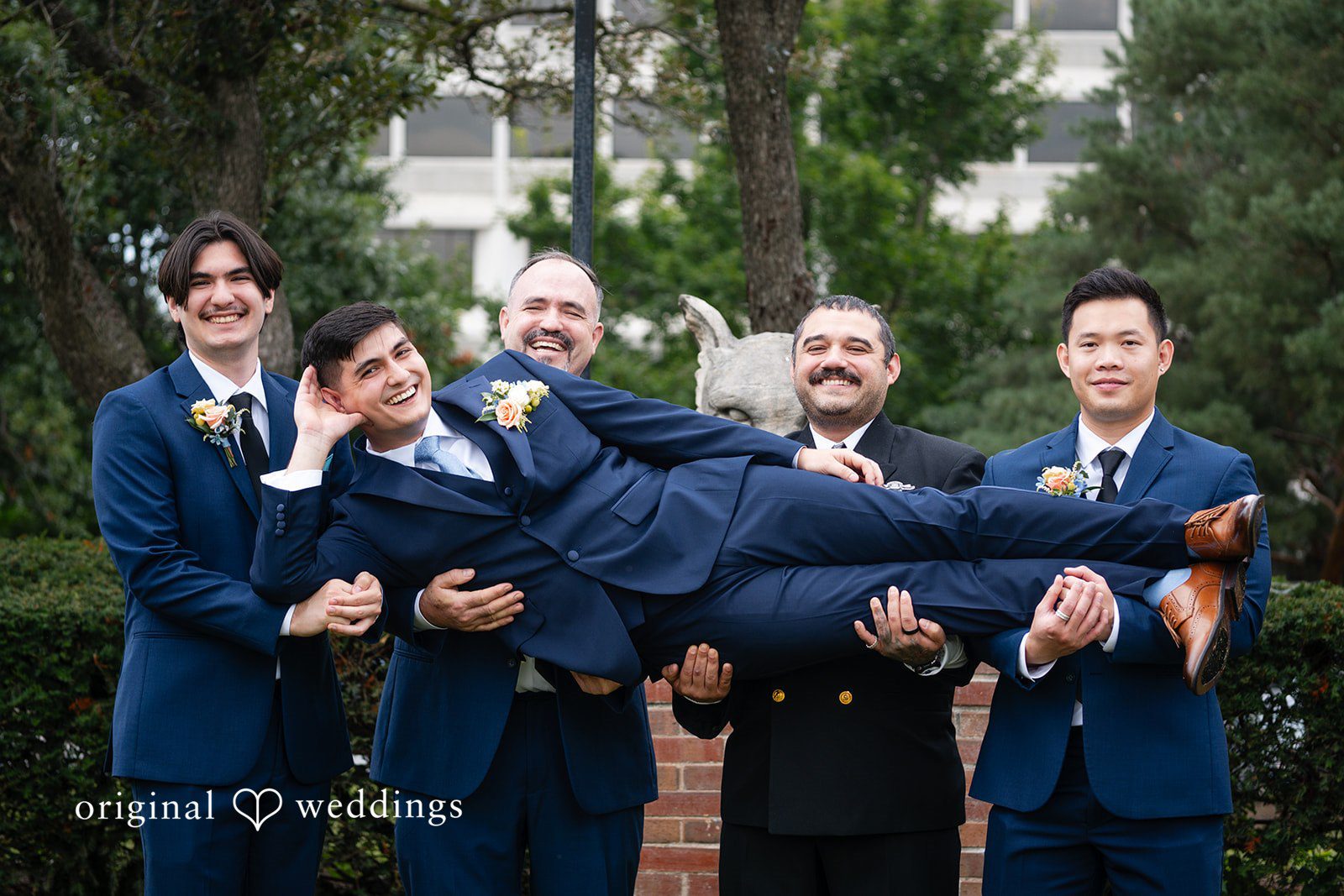 Selena + Ezequiel A group of groomsmen playfully hold up one man horizontally while posing outside.