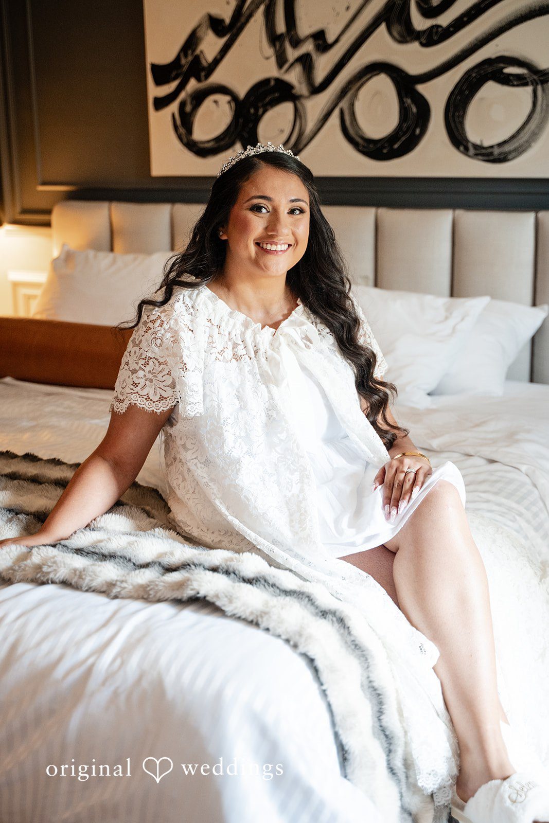 Selena + Ezequiel A woman wearing a white lace-trimmed robe sits smiling on the edge of a bed.