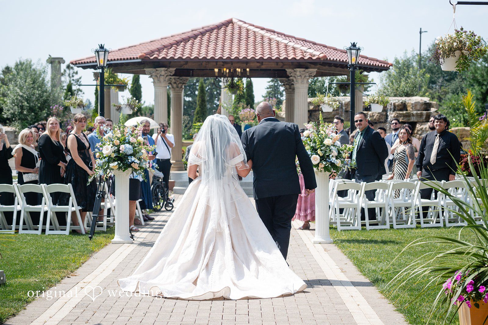 Selena + Ezequiel A bride walks down a paved aisle between rows of seated wedding guests.