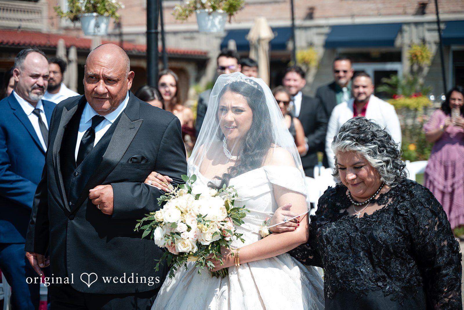 Selena + Ezequiel A bride walks arm-in-arm down an aisle with two individuals as guests look on.