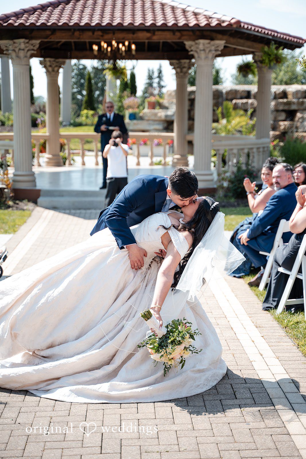 Selena + Ezequiel A bride and groom share a celebratory dip and kiss on a paved path.