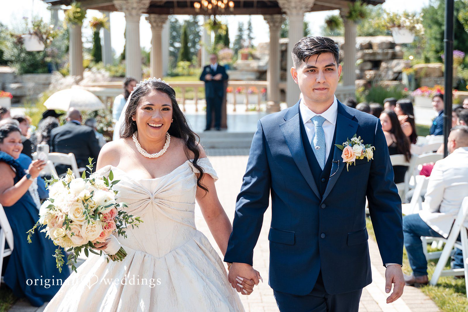 Selena + Ezequiel A bride and groom smile while walking hand-in-hand past rows of white wedding chairs.