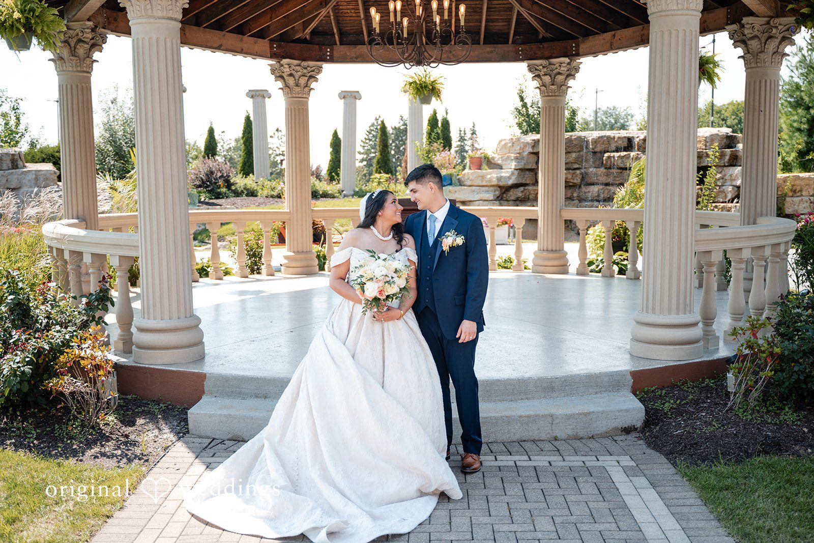 Selena + Ezequiel A bride and groom stand together posing in front of a white gazebo structure.