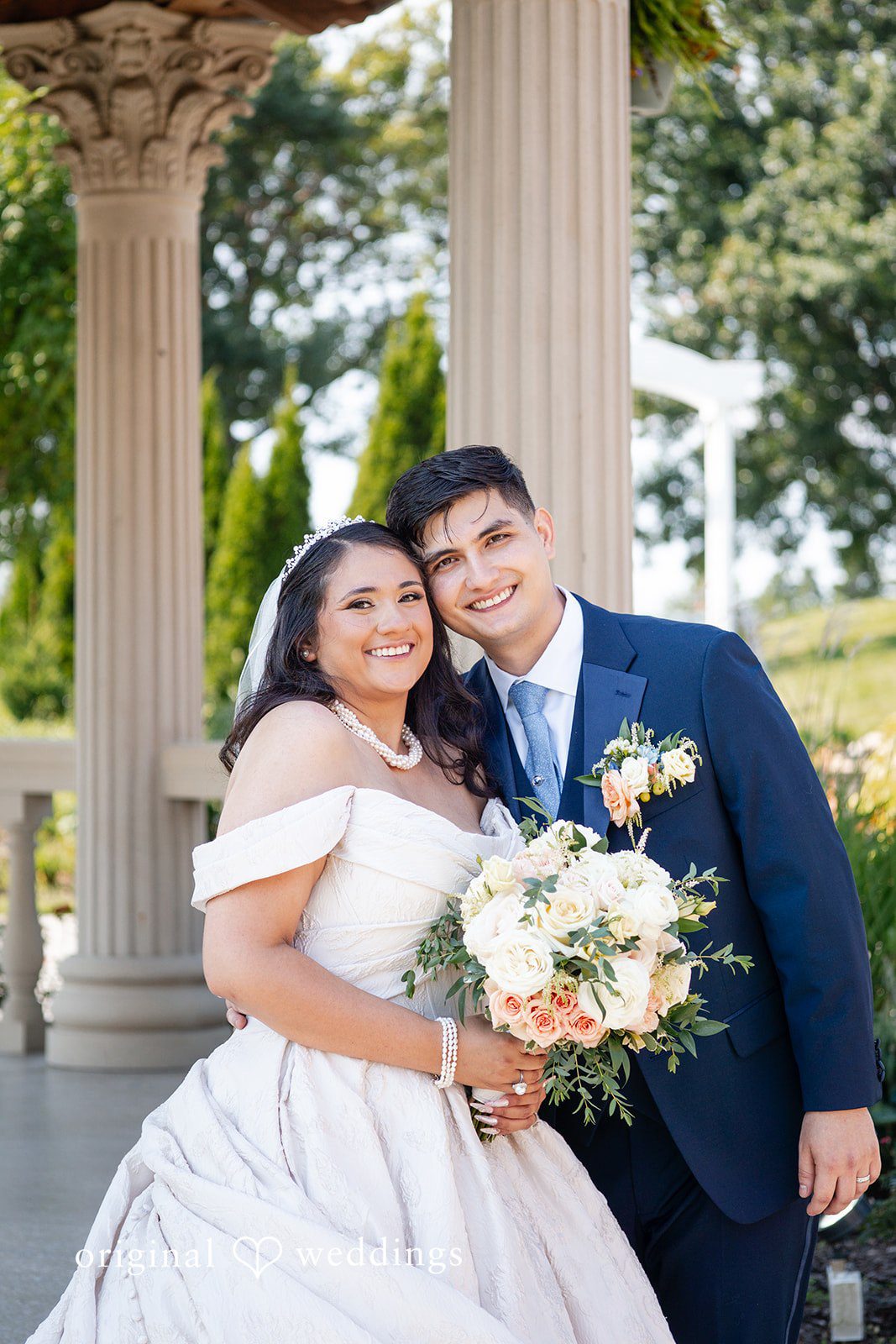 Selena + Ezequiel A bride and groom hold a white bouquet while posing closely together near greenery.