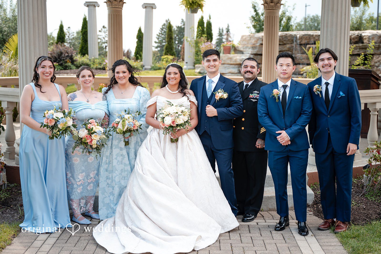 Selena + Ezequiel A wedding party stands in a line posing outdoors next to decorated stone pillars.