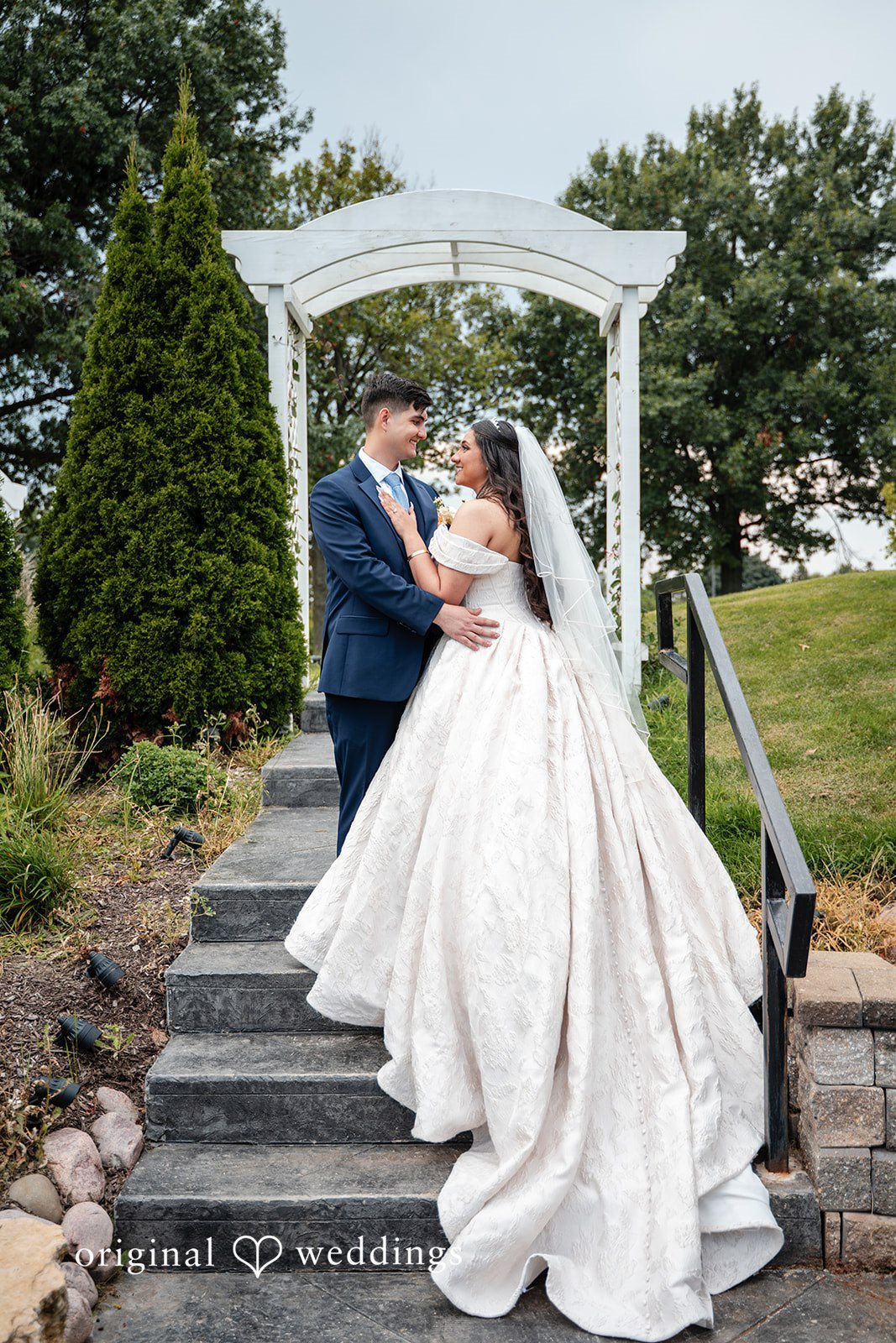 Selena + Ezequiel A bride and groom pose together on stone steps leading up toward a white structure.