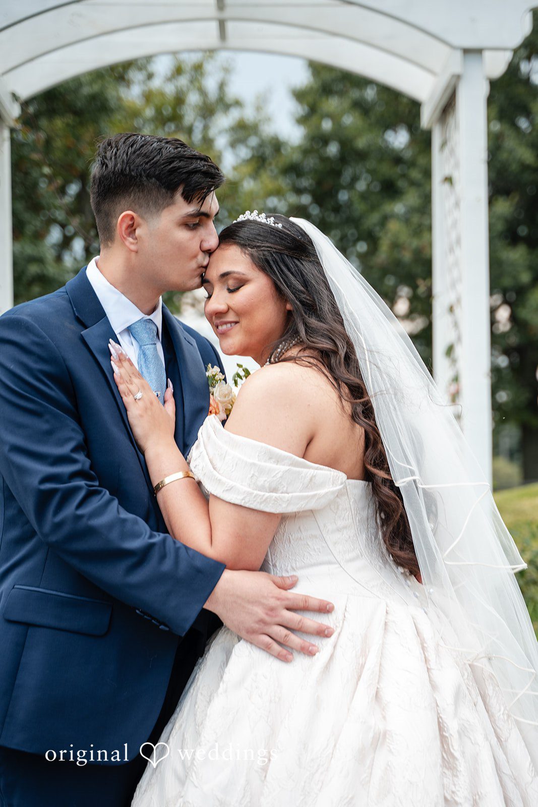 Selena + Ezequiel A groom embraces a bride from behind while standing under a white gazebo archway.