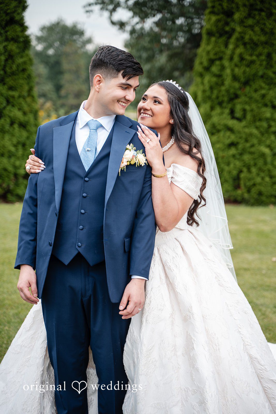 Selena + Ezequiel A bride and groom stand together on a green lawn smiling at the camera.