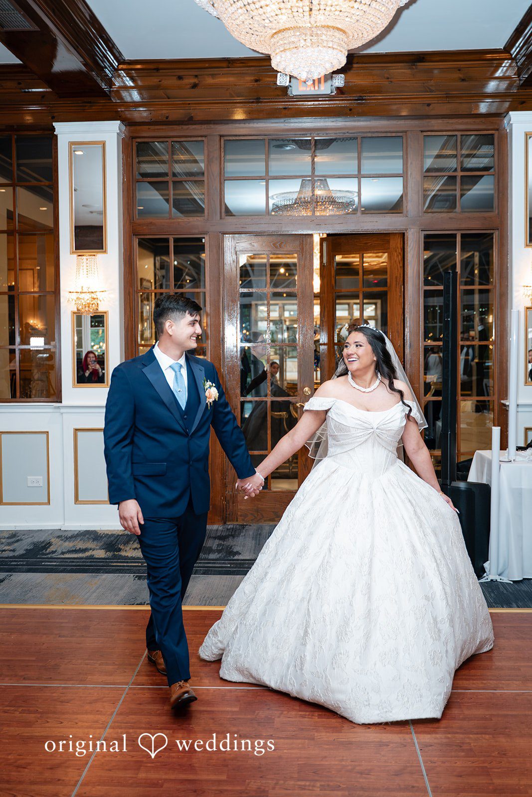 Selena + Ezequiel A bride and groom hold hands while walking together through a beautifully lit reception venue.
