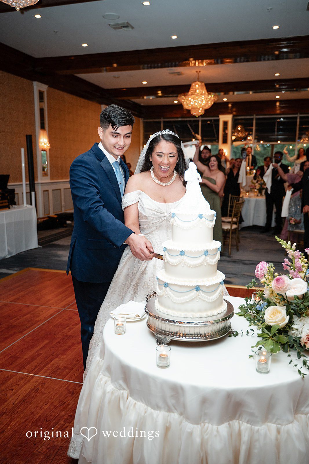 Selena + Ezequiel A bride and groom stand next to a tiered white wedding cake during their reception.