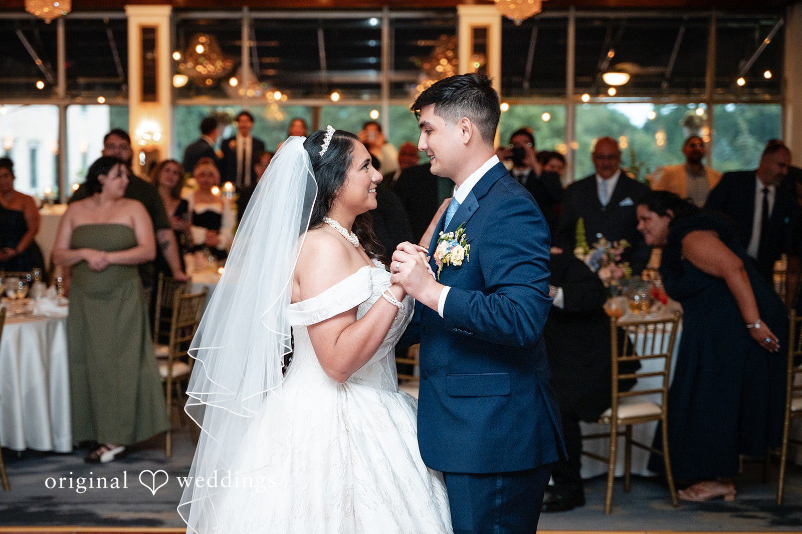 Selena + Ezequiel A bride in a white ballgown and a groom in a dark blue suit share a first dance with reception guests blurred in the background.