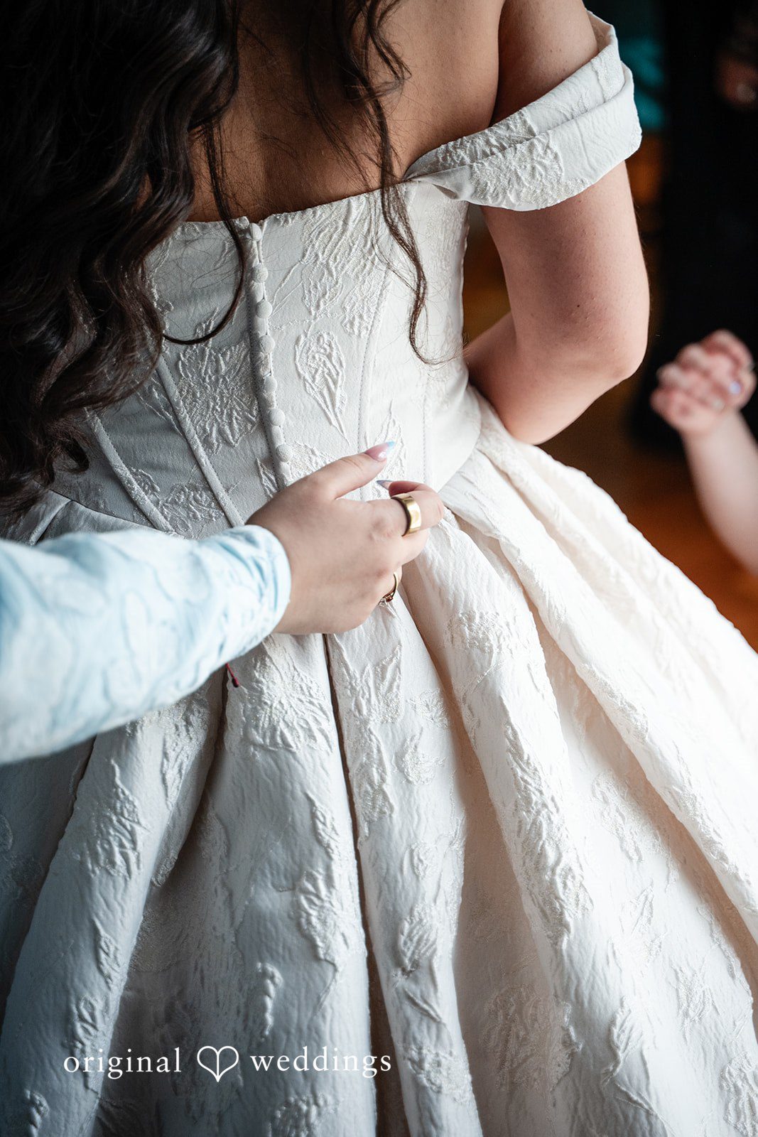 Selena + Ezequiel A close-up view of hands buttoning up the back of a white wedding gown.