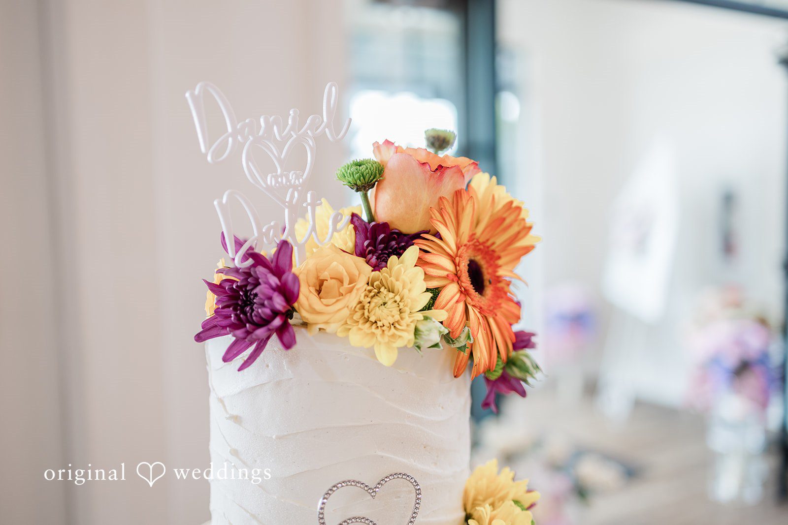 Daniel + Dalte A close-up of a white cake decorated with vibrant purple, yellow, and orange flowers.