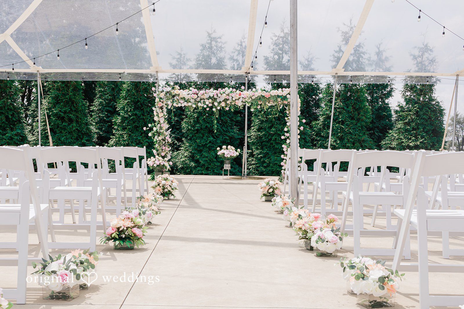Daniel + Dalte An aisle lined with white chairs leading to a wedding altar decorated with green foliage and white flowers.