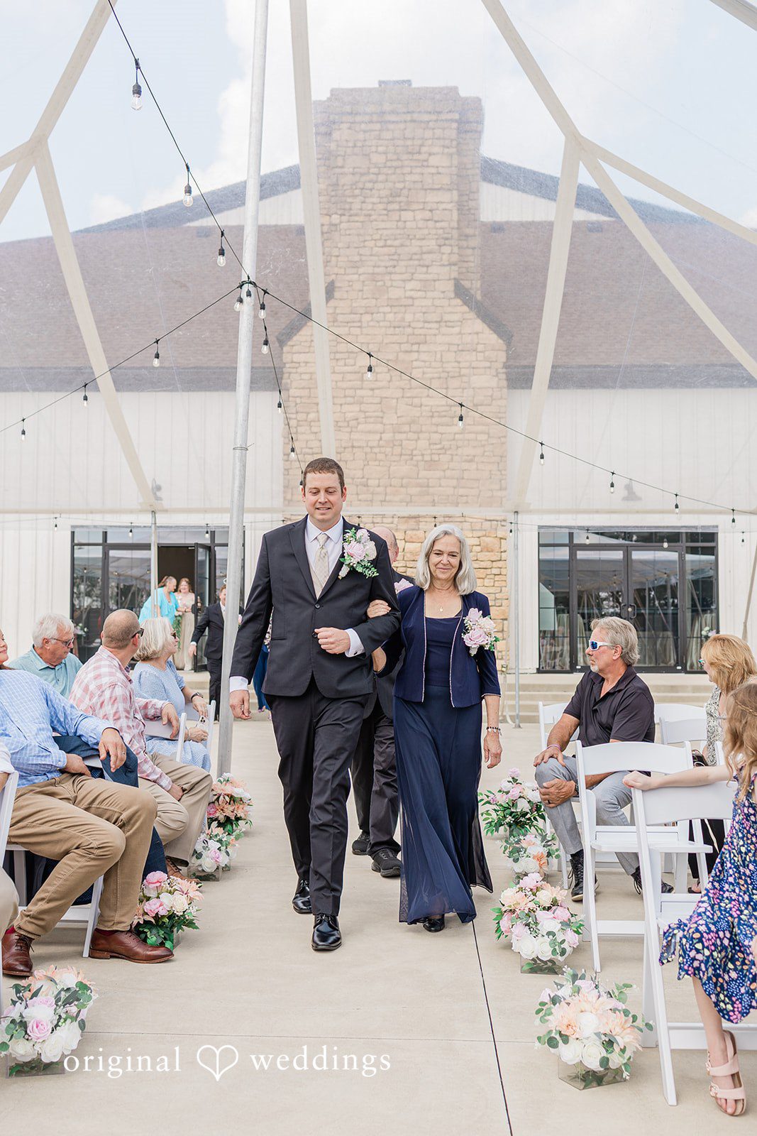 Daniel + Dalte A wedding ceremony taking place in an open structure with guests seated on white chairs.