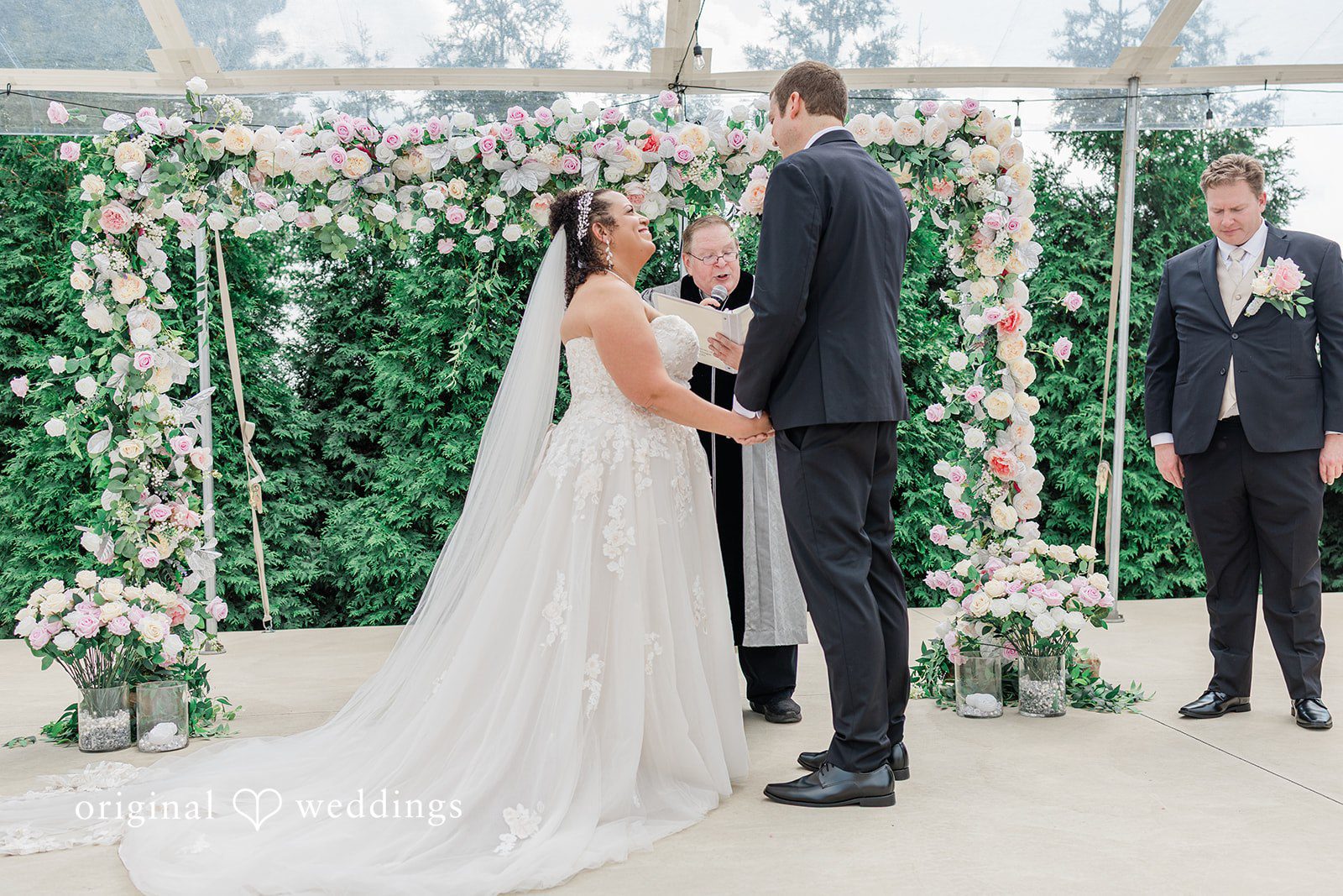 Daniel + Dalte A bride and groom stand hand-in-hand before a floral wedding arch during their ceremony.