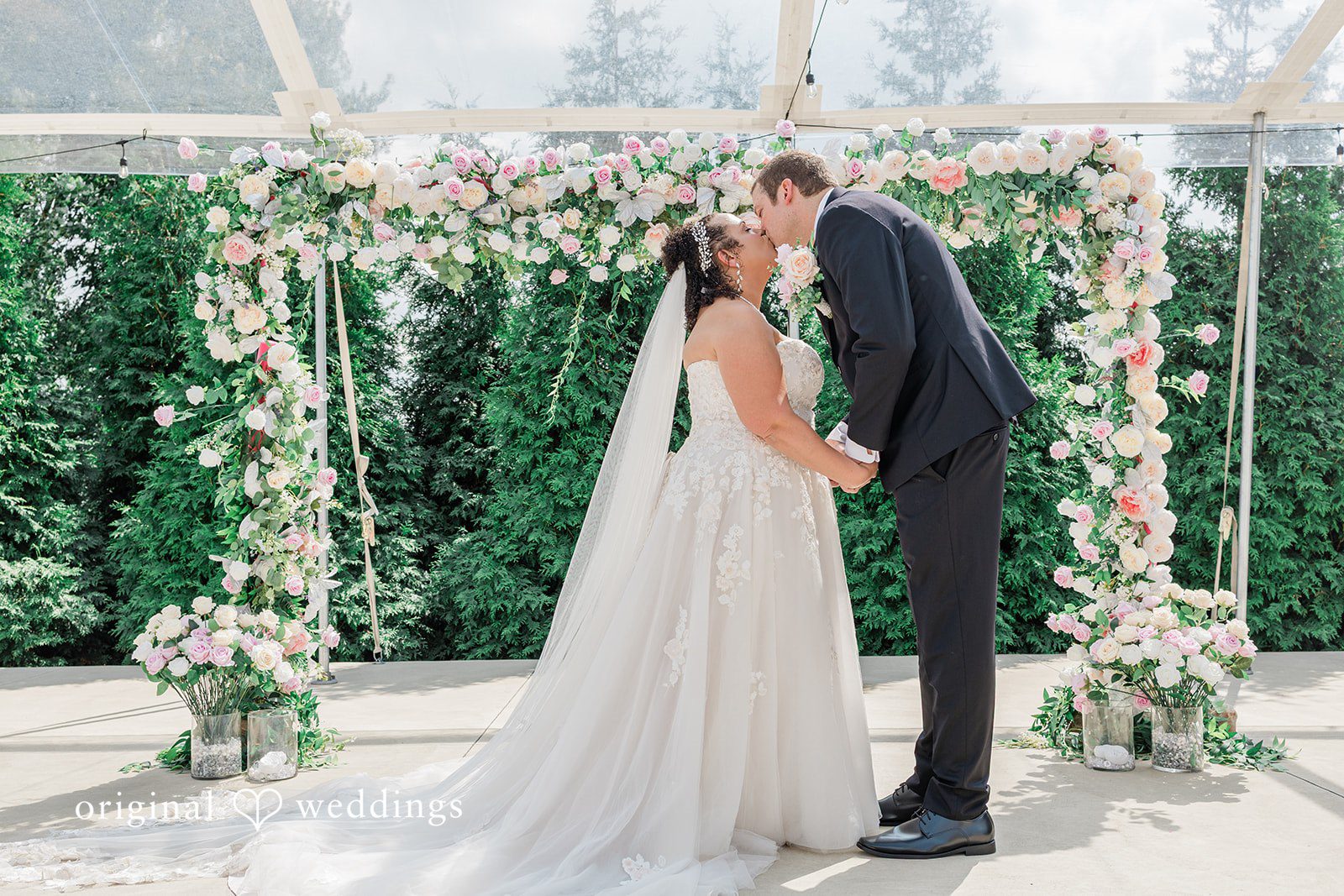 Daniel + Dalte A bride and groom share a kiss in front of a wedding arch decorated with white flowers.