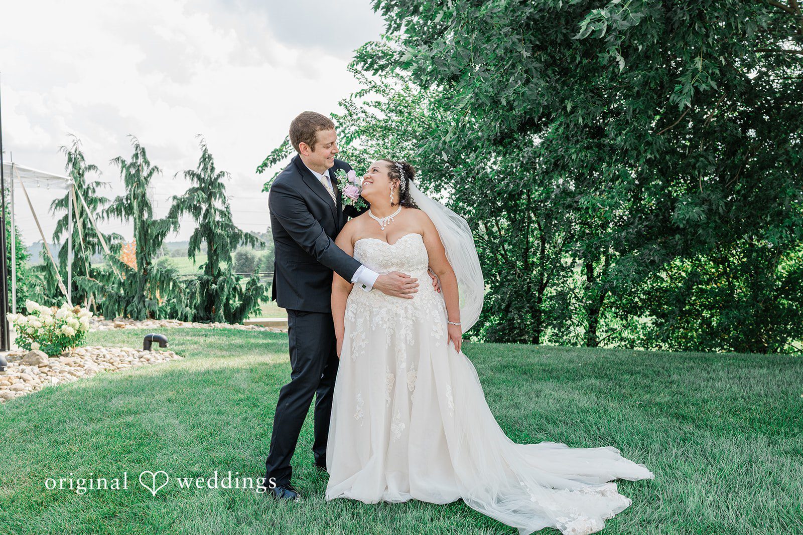 Daniel + Dalte A bride and groom embrace while standing on a grassy field with dense woods in the background.