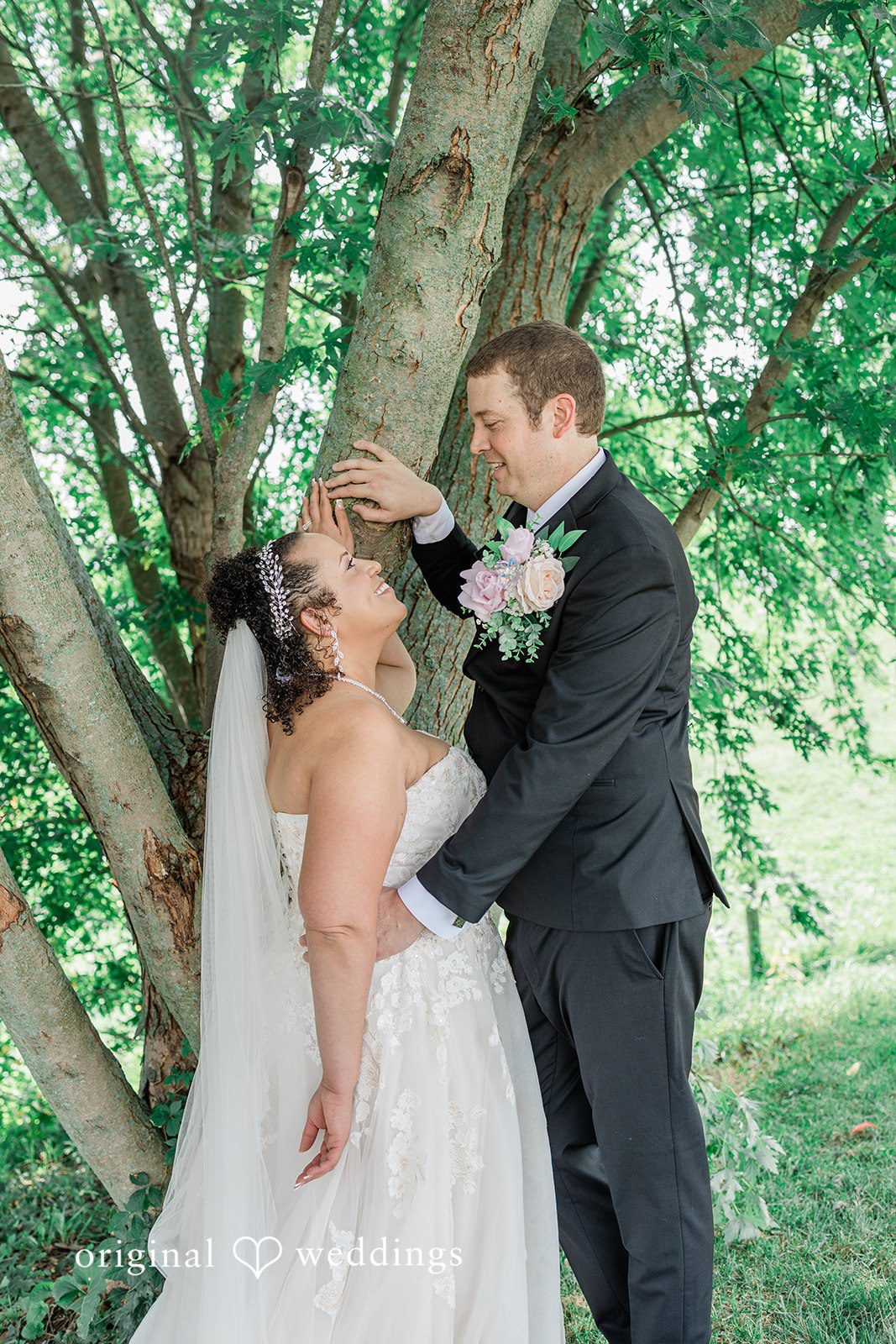 Daniel + Dalte A bride in a white gown and a groom in a black suit smiling at each other beneath a large tree.