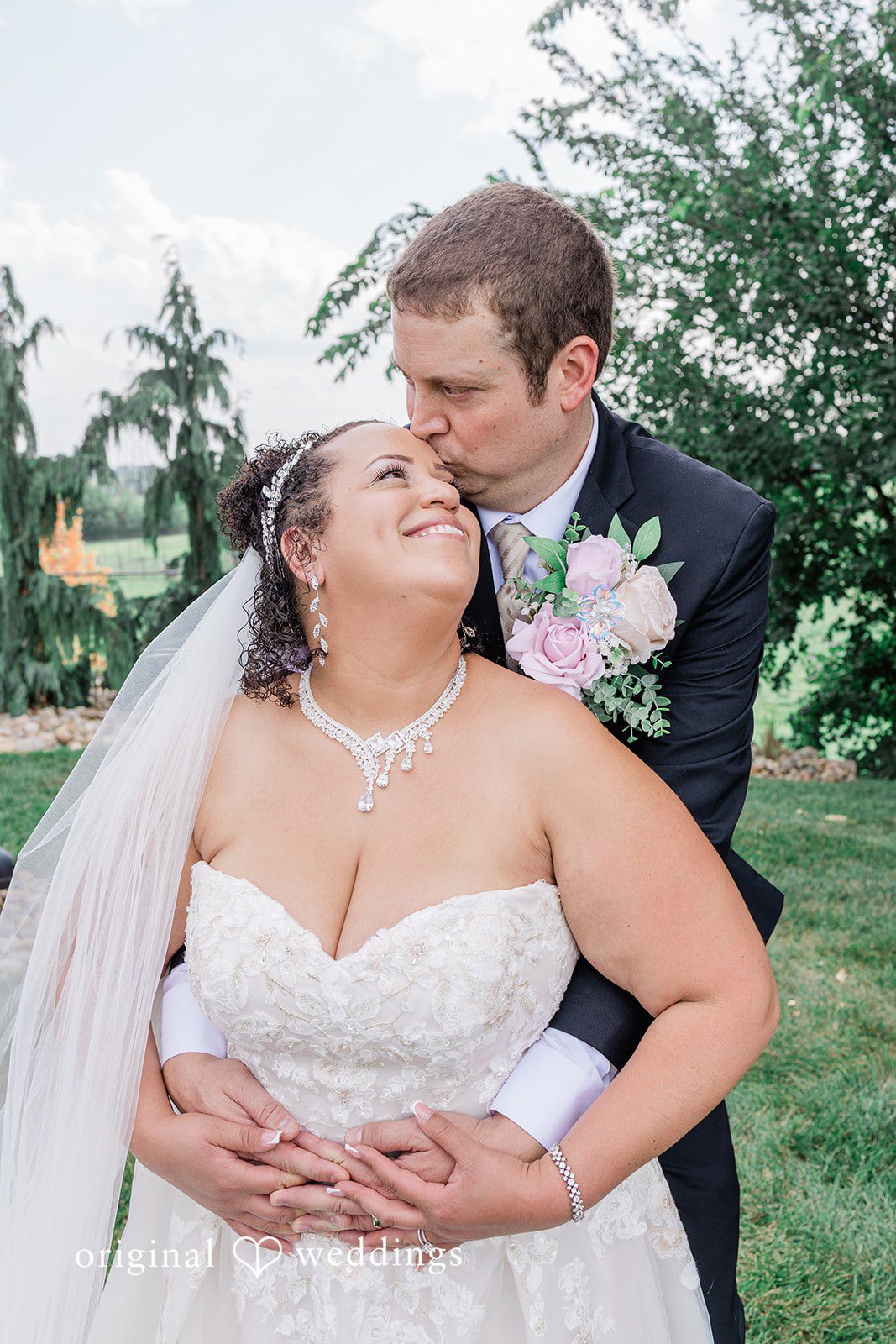 Daniel + Dalte A groom wraps his arms around a smiling bride from behind while standing on a lawn.