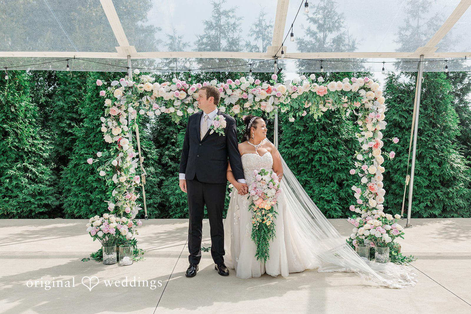 Daniel + Dalte A bride and groom pose together beneath a large archway covered in greenery and white blossoms.