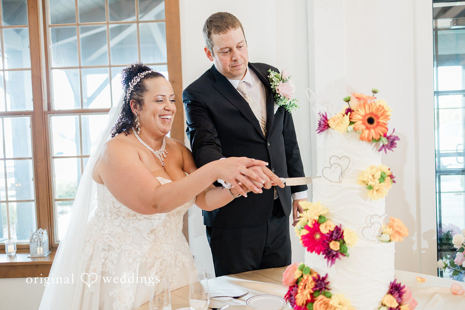 Daniel + Dalte A bride and groom smiling as they cut into a wedding cake decorated with colorful flowers.