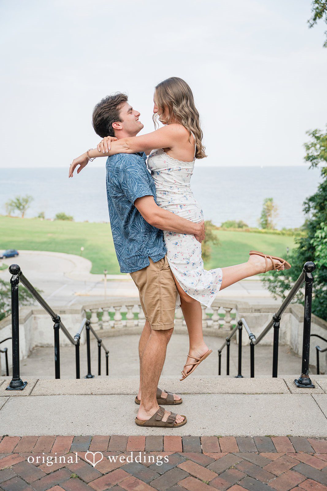 A couple laughing as one person lifts the other into the air near a waterfront walkway.