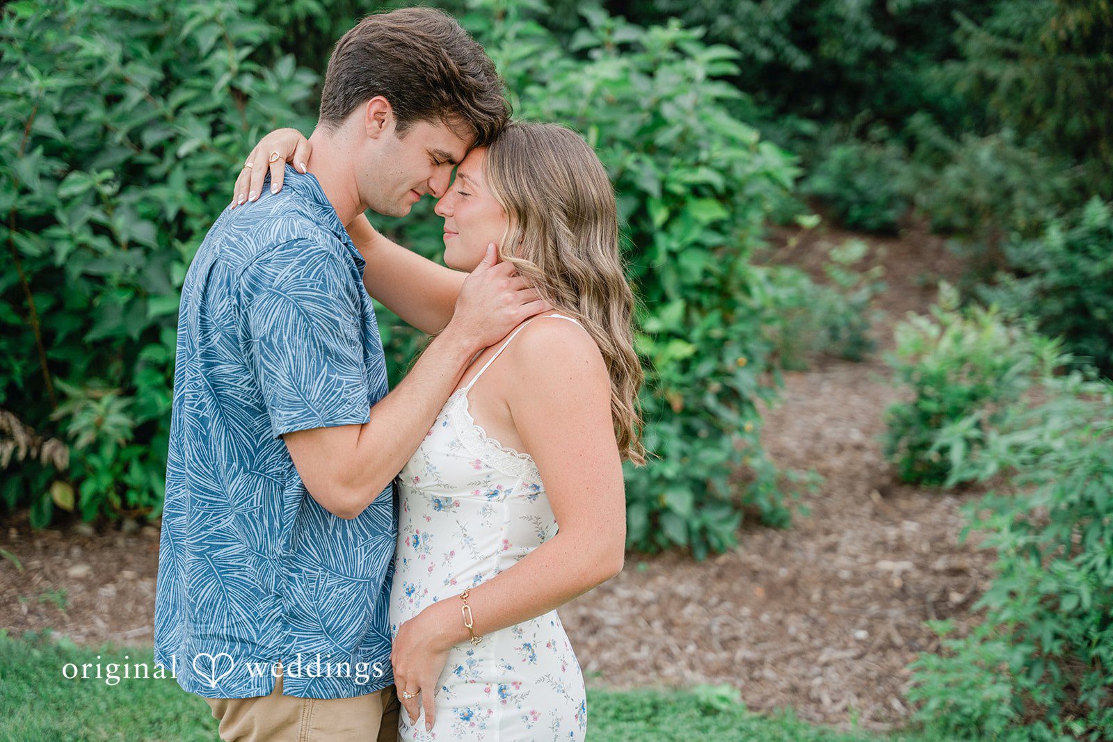 A couple facing each other smiling in front of a green leafy backdrop.