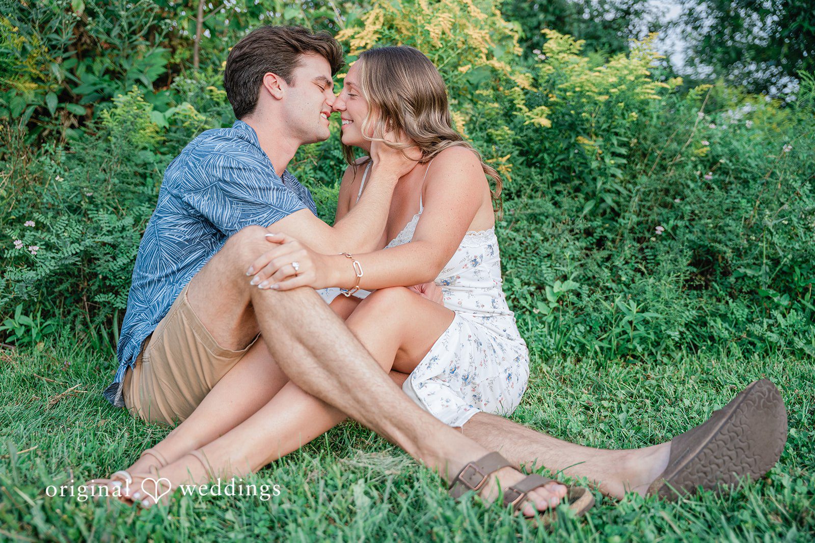 A couple sitting together on a grassy area, looking at one another.
