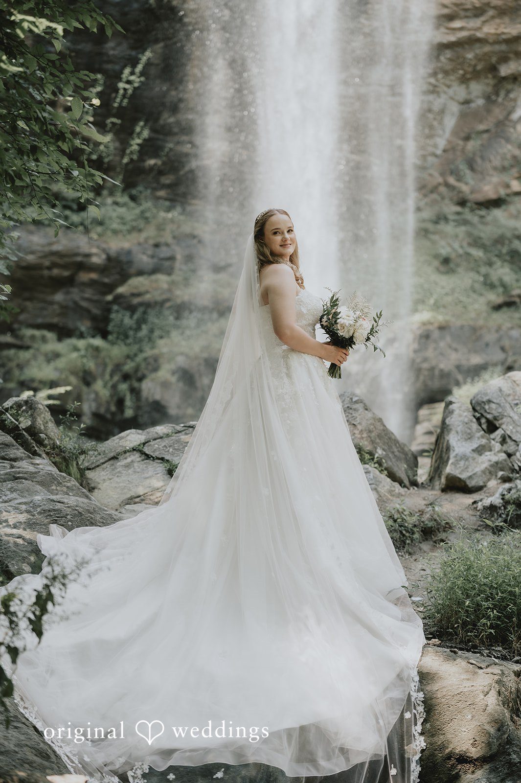 Sean + Hannah A bride with a long flowing train poses in front of a large waterfall.