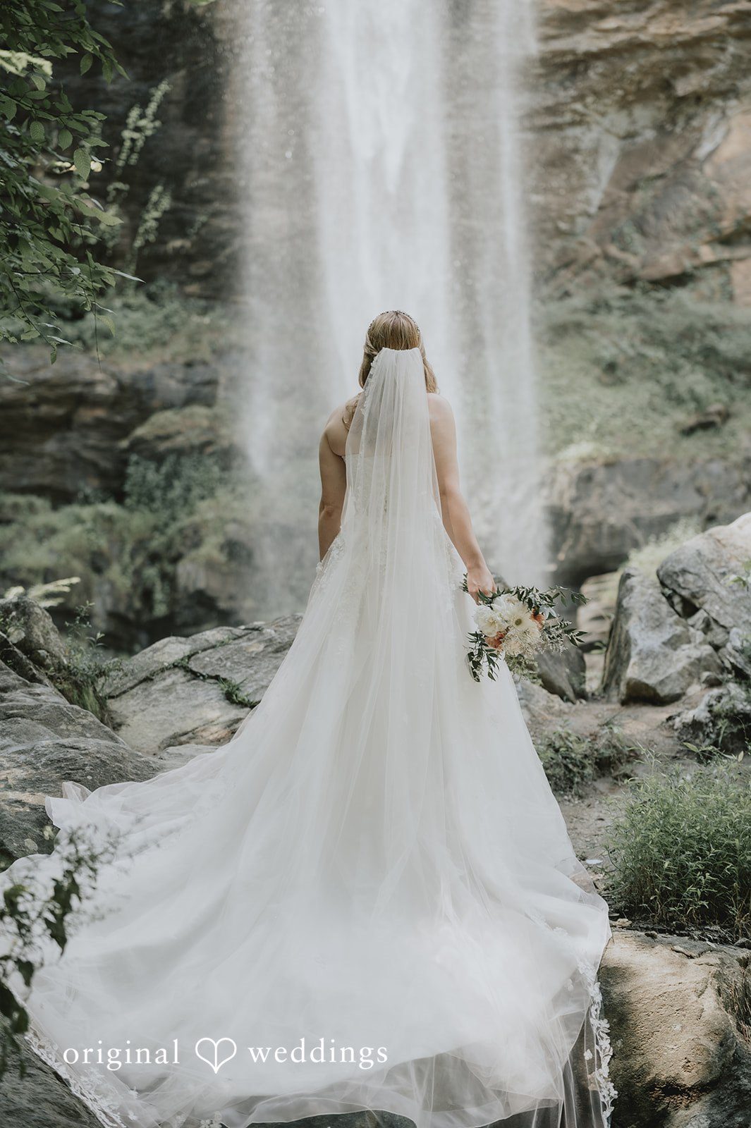Sean + Hannah A view from behind of a bride in a white wedding dress looking toward a tall waterfall.