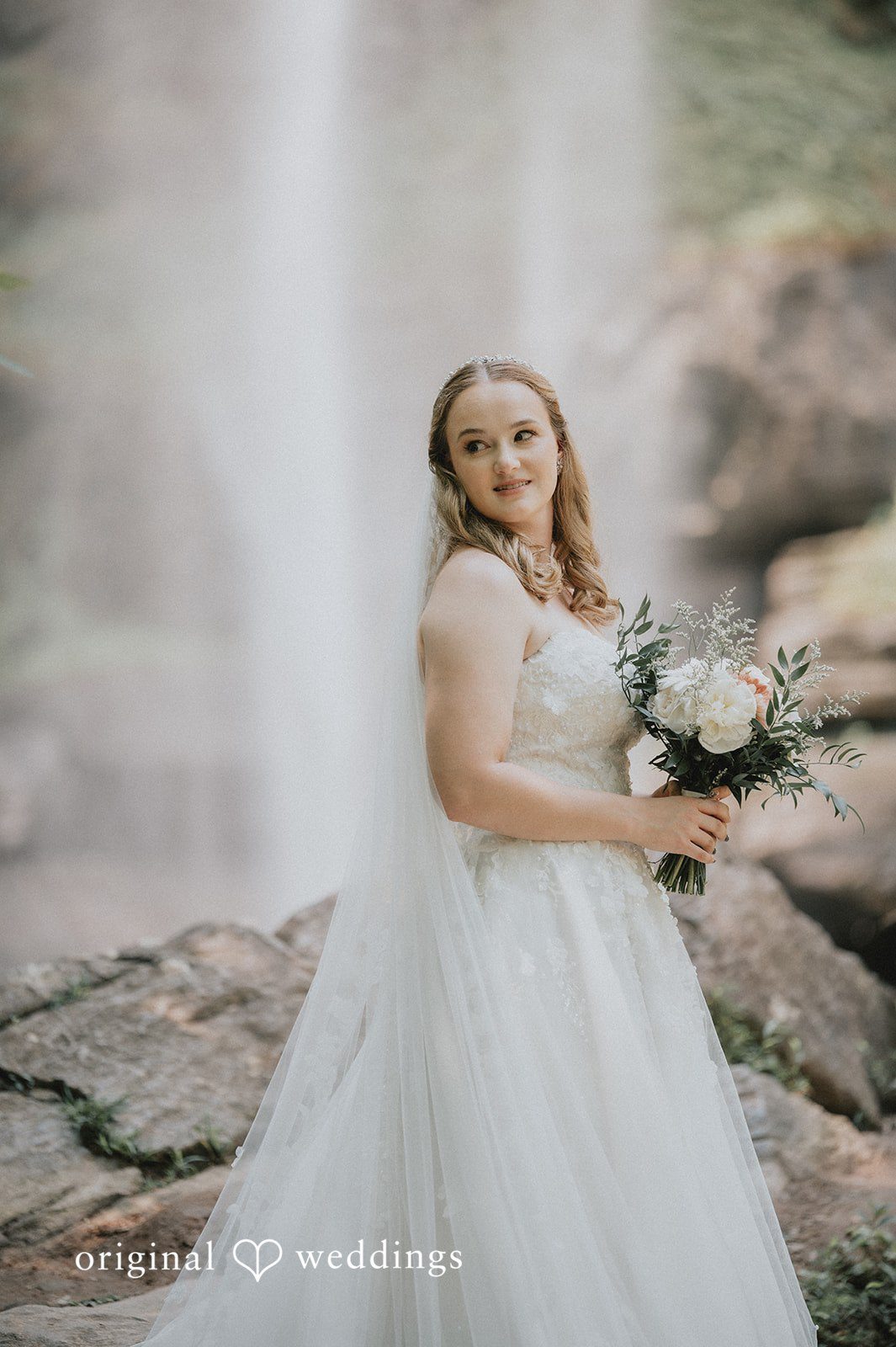 Sean + Hannah A bride in a white wedding gown holding a bouquet stands in front of a rocky background.