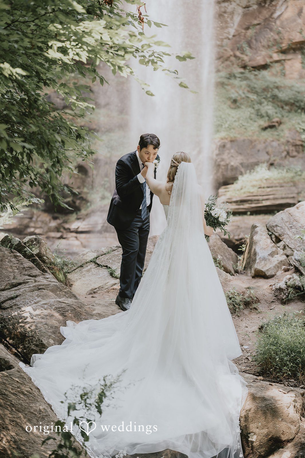 Sean + Hannah A man in a dark suit dips a woman in a flowing white dress back for a kiss at the base of a waterfall.