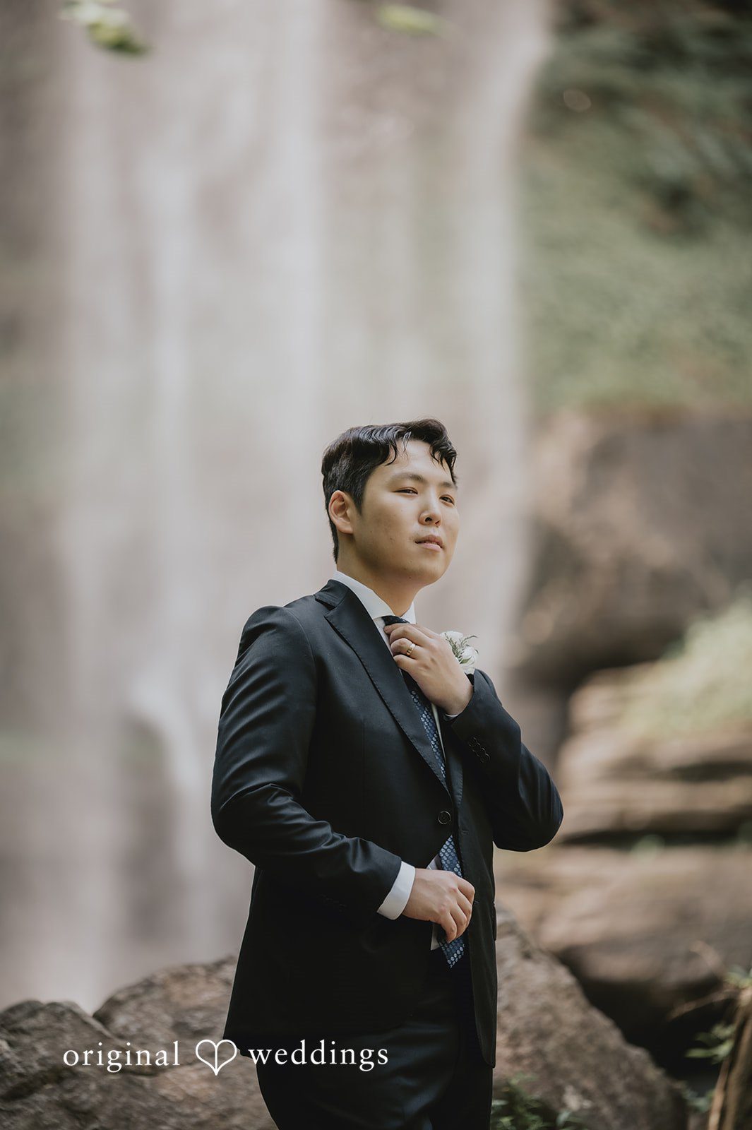 Sean + Hannah A groom in a black tuxedo stands posing with a rocky waterfall background.