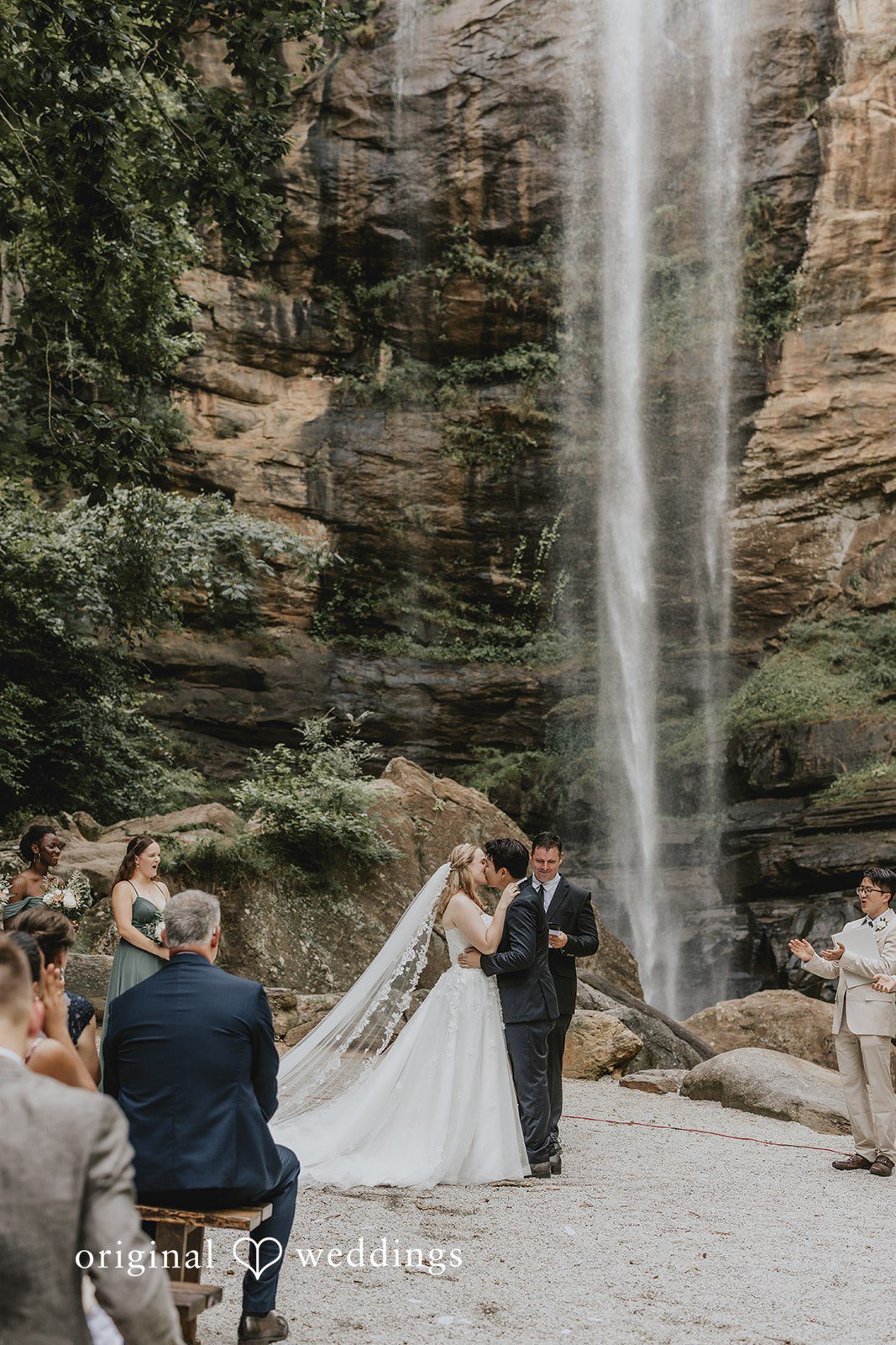 Sean + Hannah A wide-angle view of a wedding ceremony taking place at the base of a tall waterfall.