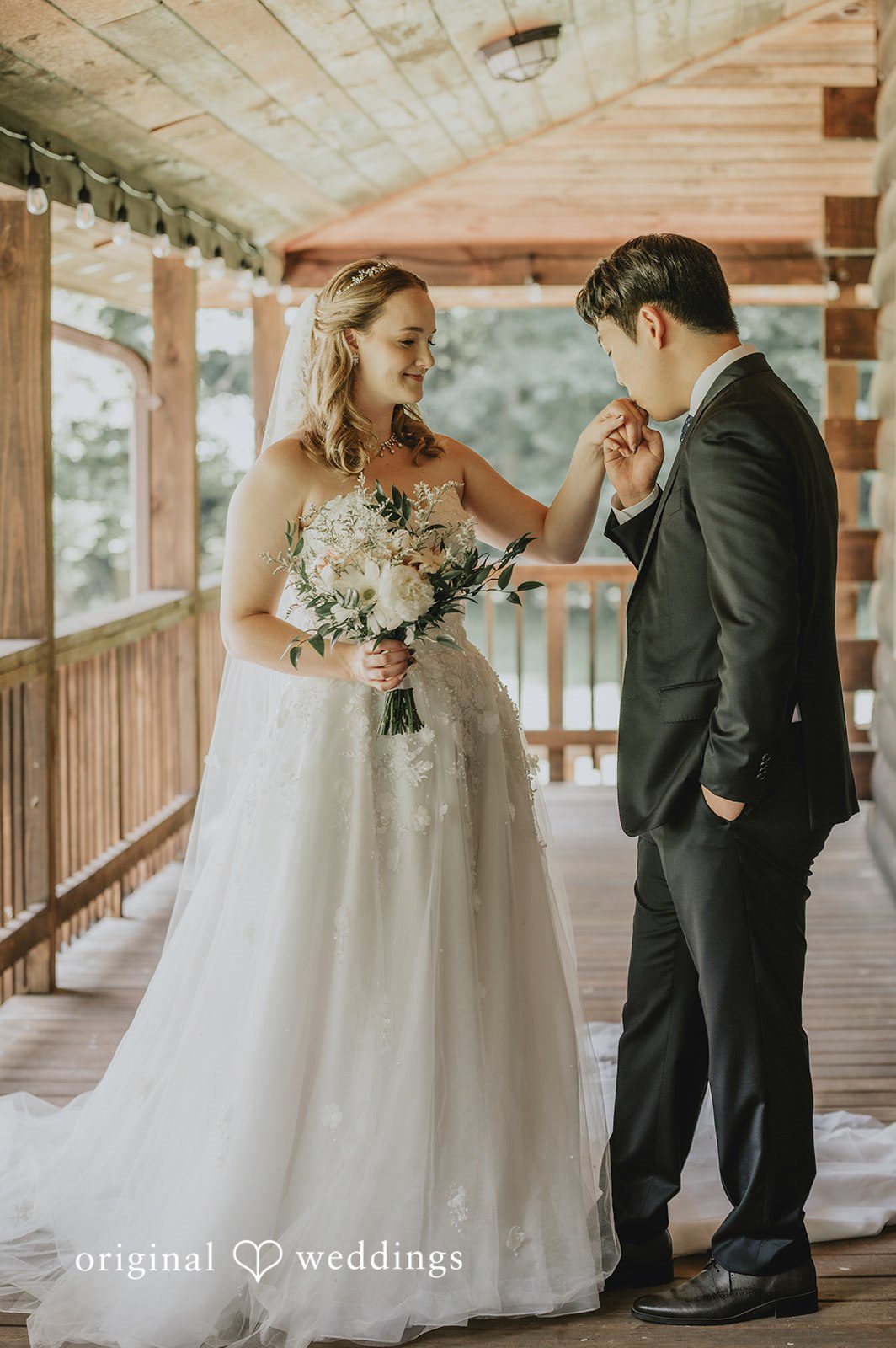 Sean + Hannah A man in a dark suit leans forward to kiss the hand of a smiling woman in a white gown on a wooden balcony.