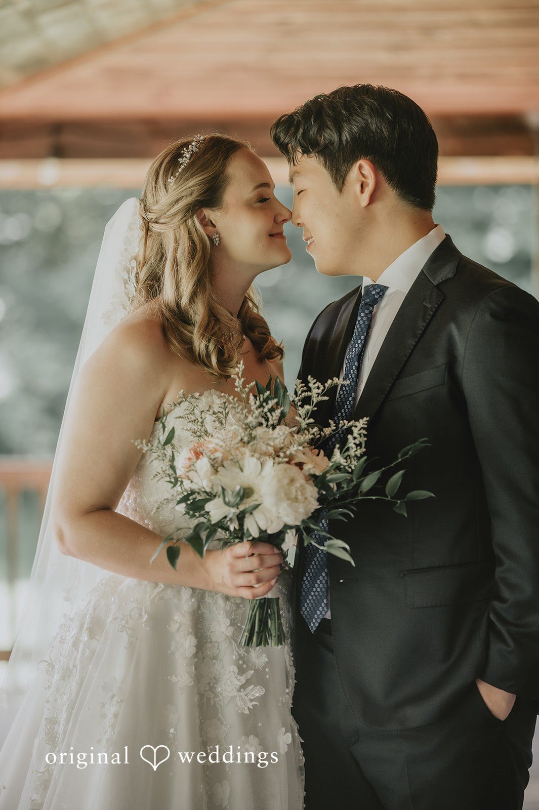 Sean + Hannah A man in a dark suit and a woman in a white lace dress smile at one another while sharing a close embrace.