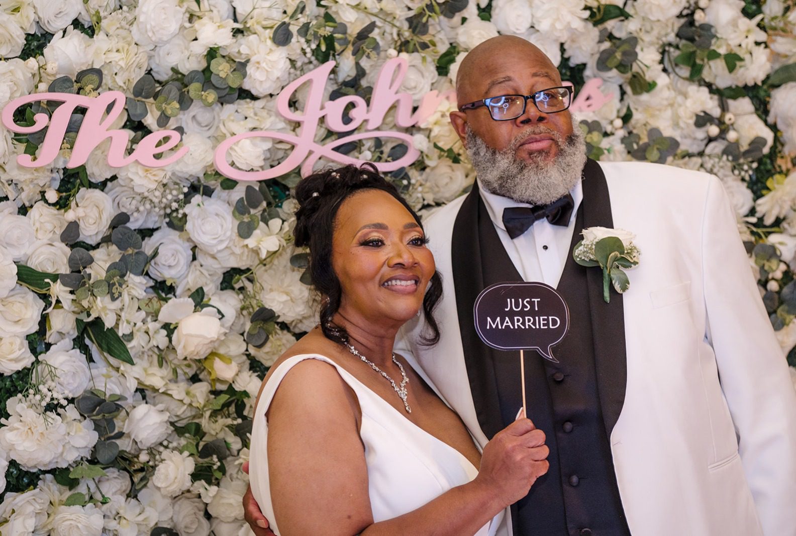 Theodore and Kimberle smiling in front of a white flower wall, captured by Cleveland wedding photographers from Original Weddings at Tom's Country Place