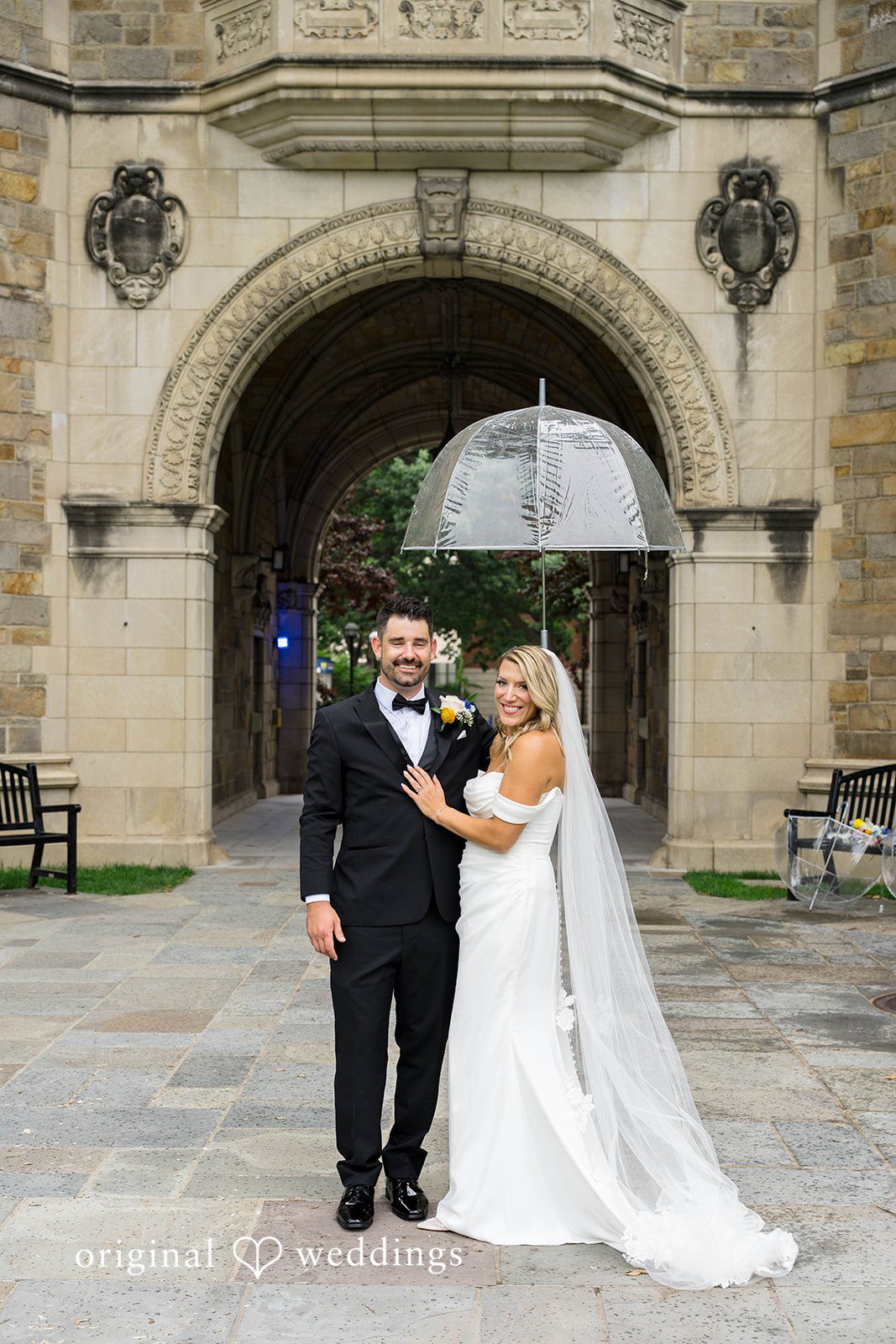 Bride and groom under a clear umbrella in a stone courtyard