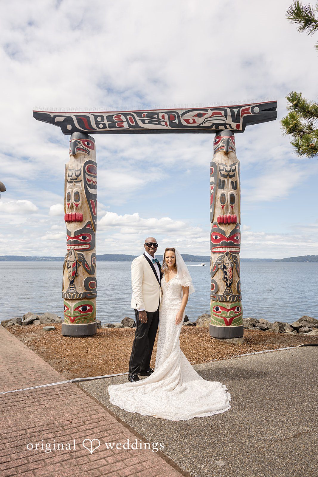 Jennifer + Tony A distant shot of a bride and groom posing under a wooden arbor by the water.