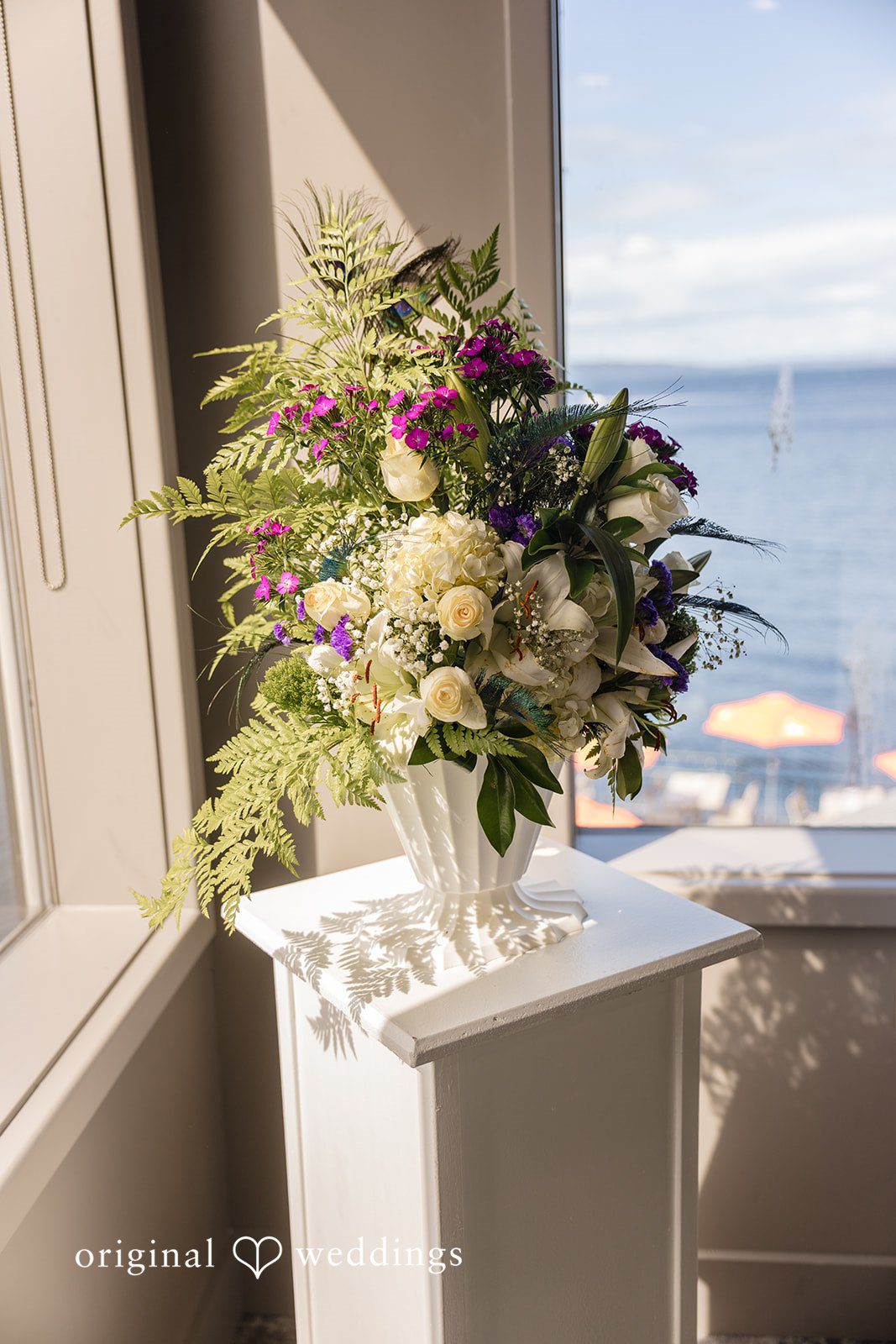 Jennifer + Tony A colorful, lush floral arrangement on a white pedestal with water in the background.