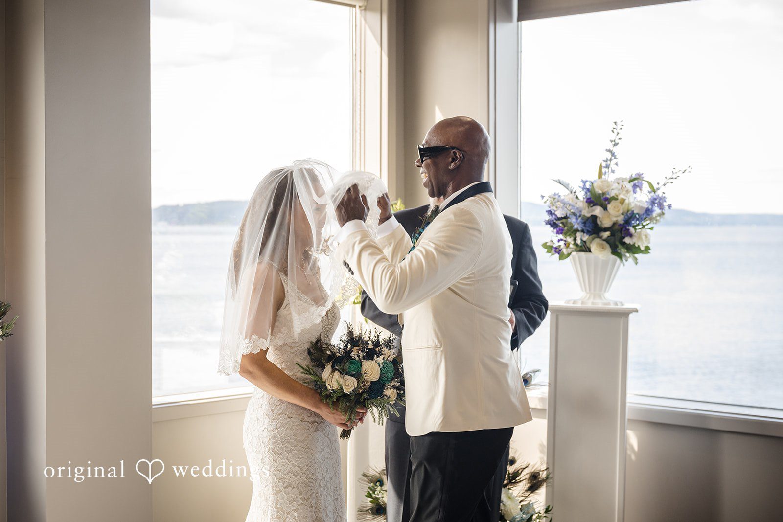 Jennifer + Tony A groom wearing a white tuxedo jacket and sunglasses gently lifts the veil off his bride's face during their wedding ceremony.