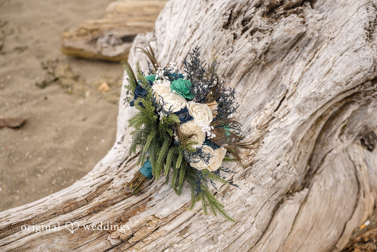 Jennifer + Tony A wedding bouquet featuring white flowers, green eucalyptus leaves, and a teal ribbon resting on a rustic, textured piece of wood.